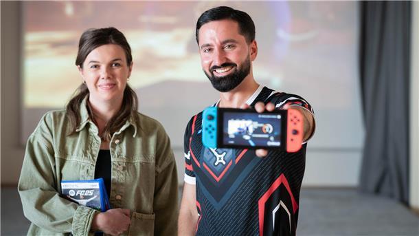 Eine Frau und ein Mann stehen nebeneinander. Der Mann hält ein Nintendo-Switch-Handheld in die Kamera, die Frau trägt zwei Spielhüllen im Arm.