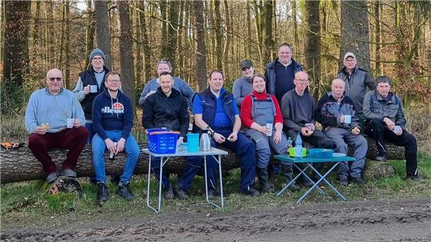 Eine Gruppe von Leuten sitzt auf einem Baumstamm. Sie trinken Kaffee und lächeln in die Kamera.