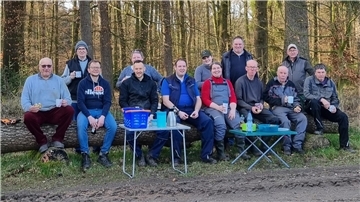 Eine Gruppe von Leuten sitzt auf einem Baumstamm. Sie trinken Kaffee und lächeln in die Kamera.