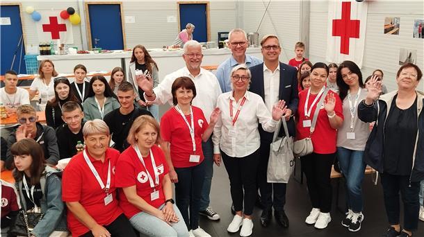 Eine Gruppe von Kindern, Jugendlichen und Erwachsenen winken in die Kamera. Im Hintergrund hängen Flaggen mit dem Symbol des Roten Kreuzes.