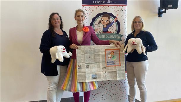 Zauberhafte Zahnpflege: Kinder-Show in der Grundschule Harrien Drei Frauen stehen nebeneinander und präsentieren Zahn-Stofftiere sowie eine aufgeschlagene Zeitung.