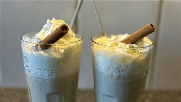 Zwei Gläser mit Latte und einer Zimtstange als Deko stehen auf einer Arbeitsfläche in einer Küche.