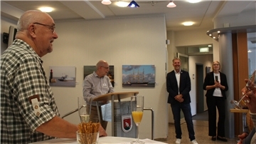 Bei einer Ausstellungseröffnung im Rathaus Beverstedt spricht ein Mann am Rednerpult. Gäste hören zu, einige halten Sektgläser in den Händen. An den Wänden hängen Fotografien mit maritimen Motiven
