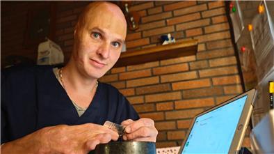 Ein Mann mit Brandnarben im Gesicht sitzt an einem Tisch vor einem Ipad. In seiner Hand hält er einen Tablettenblister, den er über einen Mörser hält. 