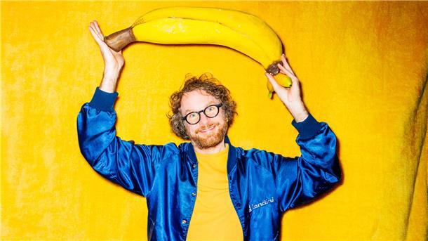 Ein Brille tragender Mann in einem gelben Hemd und blauer Jacke steht vor einen gelben Wand - und hält eiine überdimensionierte Banane in den Händen. Riesen-Banane oder Mini-Klon? Friedemann Weise stellt in seiner Show „Das bisschen Content“ eine Miniaturausgabe von sich selbst vor. Deshalb wirkt die Banane auf dem Plakat so groß.