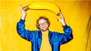 Ein Brille tragender Mann in einem gelben Hemd und blauer Jacke steht vor einen gelben Wand - und hält eiine überdimensionierte Banane in den Händen. Riesen-Banane oder Mini-Klon? Friedemann Weise stellt in seiner Show „Das bisschen Content“ eine Miniaturausgabe von sich selbst vor. Deshalb wirkt die Banane auf dem Plakat so groß.