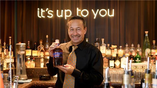 Ein Mann in einer Bar, der seine Rumkreation zeigt. Im Hintergrund ist in leuchtender Schrift "it's up to you" zu lesen. 