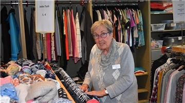 Eine ältere Frau in einem grauen Pullover steht vor einem Kleiderständer.