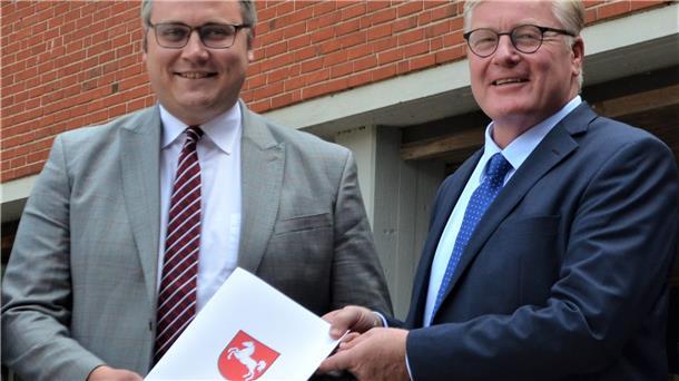 Zwei Männer halten ein Papier mit dem aufgedruckten Wappen Niedersachsens in Händen. 