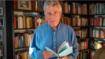 Ein älterer Mann hält ein Buch in der Hand. Er steht vor einem Bücherregal.
