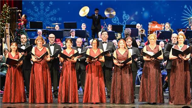 Ein Chor, bestehend aus Männern und Frauen, trägt elegante Abendkleidung. Die Frauen tragen rote, bodenlange Kleider, die Männer schwarze Anzüge mit Fliege. Sie halten Notenblätter und singen vor einem großen Orchester im Hintergrund. Der Bühnenhintergrund zeigt winterliche Motive, darunter Schneeflocken und festliche Dekorationen.