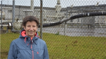 Eine Frau steht vor einem Maschendrahtzaun, der eine Gasförderstation einzäunt.