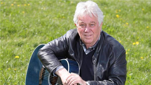 Der Sänger Rolf Zuckowski sitzt mit seiner Gitarre auf einer Wiese.