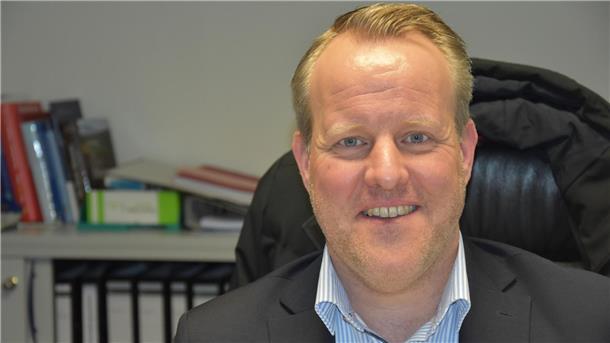 Ein blonder Mann mittleren Alters sitzt auf einem Bürostuhl und lächelt in die Kamera. Hinter ihm sind Aktenordner erkennbar.