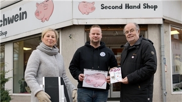 Eine Frau und zwei Männer stehen vor dem Eingang eines Geschäfts. Sie halten verschiedene Dinge in Händen, eine Klingel, ein Bild eines Sparschweins sowie eine mobile Rampe.
