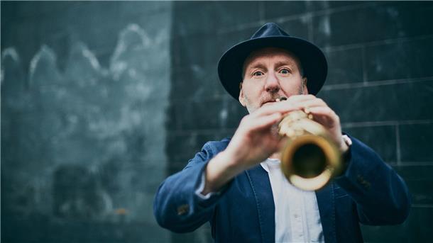 Ein Mann mit Hut spielt Saxophon und blickt direkt in die Kamera. Die Aufnahme ist leicht unscharf im Vordergrund und zeigt eine dunkle Wand im Hintergrund.