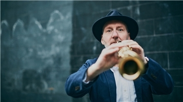 Ein Mann mit Hut spielt Saxophon und blickt direkt in die Kamera. Die Aufnahme ist leicht unscharf im Vordergrund und zeigt eine dunkle Wand im Hintergrund.