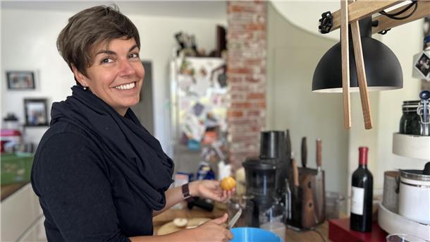 Eine Frau mit kurzen Haaren, dunkler Kleidung und Schal steht in ihrer Küche und schneidet Quitten.