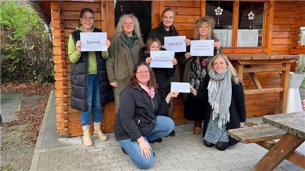 Sechs Frauen und ein Mädchen stehen und knien vor einer Holzhütte. Sie halten Zettel und einen Umschlag in die Kamera.