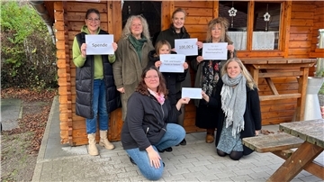 Sechs Frauen und ein Mädchen stehen und knien vor einer Holzhütte. Sie halten Zettel und einen Umschlag in die Kamera.