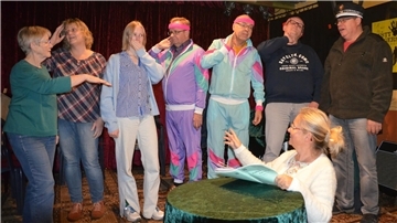 Eine bunt kostümierte Gruppe von sieben Personen steht auf einer Theaterbühne. Eine Frau mit Brille sitzt im Vordergrund an einem Tisch, hält ein Skript und gibt offensichtlich Regieanweisungen. 