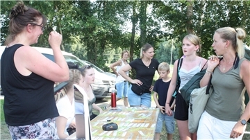 Eltern und Kinder stehen an einem Anmeldetisch im Freien, umgeben von Bäumen. Auf dem Tisch liegen Namensschilder, im Hintergrund sind Erwachsene und Fahrzeuge zu sehen.