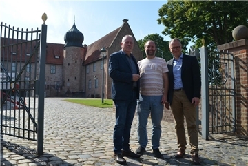 Drei Männer stehen am geöffneten Burgtor der Burg Bederkesa, im Hintergrund das historische Backsteingebäude mit Zwiebelturm bei sonnigem Wetter.