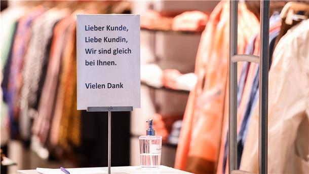 Am Eingang eines Bekleidungsgeschäftes steht ein Desinfektionsmittelspender und ein Schild mit der Aufschrift "Lieber Kunde, Liebe Kundin, wir sind gleich bei Ihnen. Vielen Dank".