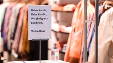 Am Eingang eines Bekleidungsgeschäftes steht ein Desinfektionsmittelspender und ein Schild mit der Aufschrift "Lieber Kunde, Liebe Kundin, wir sind gleich bei Ihnen. Vielen Dank".