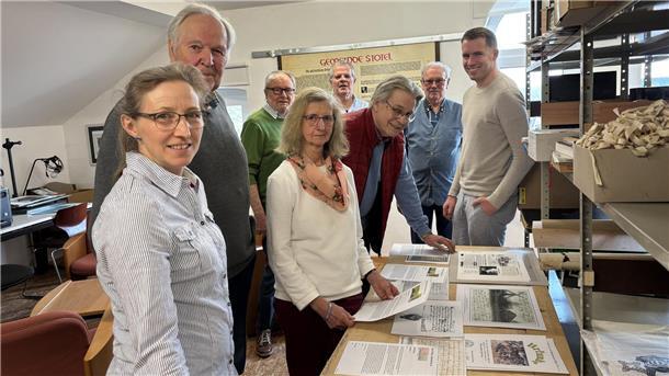 Eine Gruppe von acht Personen steht in einem Archivraum um einen Tisch mit historischen Dokumenten. Sie blicken in die Kamera, einige lächeln. Im Hintergrund sind Regale mit Akten und ein großes Informationsschild zur Geschichte von Stotel zu sehen.