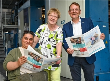 Zwei Frauen und ein Mann mit Zeitungen in der Hand. Eine Frau sitzt im Rollstuhl.