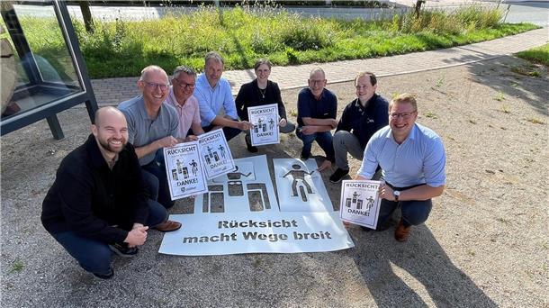 8 lächelnde Leute sitzen in der Hocke mit Plakaten „Rücksicht macht Wege breit“