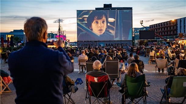 Im Fischereihafen ist eine riesige Leinwand aufgebaut, auf der ein Film zu sehen ist. Davor sitzen Hunderte Besucher. 