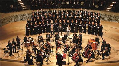 Großes Oratorienkonzert mit Chor und Orchester auf einer Konzertbühne. Im Vordergrund spielt ein Ensemble auf historischen Instrumenten, dahinter singt ein großer gemischter Chor in mehreren Reihen. Der Dirigent steht in der Mitte mit dem Rücken zum Publikum.