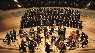 Großes Oratorienkonzert mit Chor und Orchester auf einer Konzertbühne. Im Vordergrund spielt ein Ensemble auf historischen Instrumenten, dahinter singt ein großer gemischter Chor in mehreren Reihen. Der Dirigent steht in der Mitte mit dem Rücken zum Publikum.