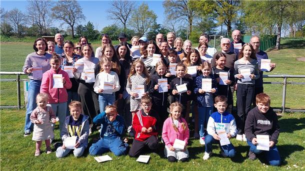 TSV Bramel ehrt 63 Sportabzeichen-Gewinner aller Altersklassen Gruppenfoto vor einem Sportplatz: etwa 30 Teilnehmende aller Altersstufen von Kleinkindern bis Senioren stehen bei sonnigem Wetter auf einer Wiese und halten stolz ihre Urkunden für das Deutsche Sportabzeichen in den Händen.