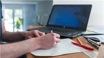 Ein Laptop steht im Hintergrund, im Vordergrund sieht man Hände Notizen auf einem Blatt Papier machen, Stifte, Geodreieck und Kalender liegen auf dem Tisch.