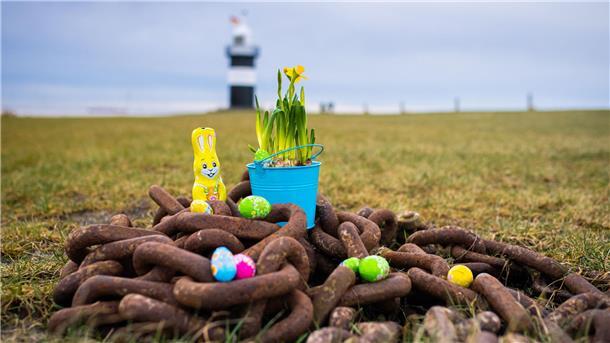 Im Vordergrund liegt eine verrostete schwere Kette, darauf liegt Osterdeko. Im Hintergrund der Leuchtturm "Kleiner Preuße" in Wremen.