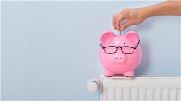 Ein Sparschwein mit Brille steht auf einer Heizung. Eine Hand lässt ein Geldstück hineinfallen.
