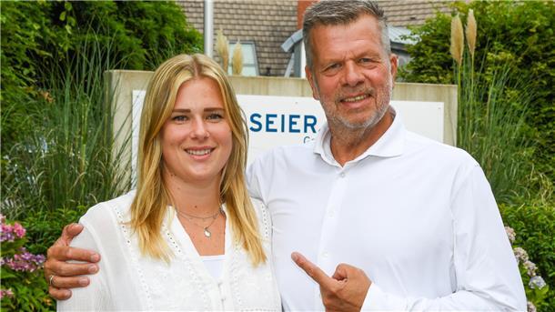 eine junge Frau und ein älterer Mann in weißer Bekleidung vor einem Firmenschild zwischen Pflanzen