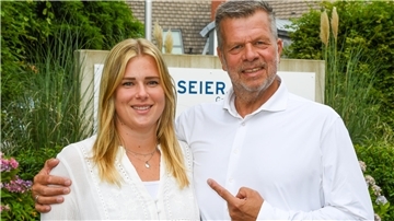 eine junge Frau und ein älterer Mann in weißer Bekleidung vor einem Firmenschild zwischen Pflanzen
