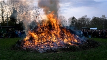 Großes Osterfeuer aus brennenden Ästen und Zweigen auf einer Wiese, umgeben von zahlreichen Zuschauerinnen und Zuschauern im Hintergrund; dichter Rauch steigt in den abendlichen Himmel, während die Flammen hell lodern.
