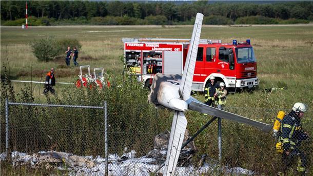 Trümmerteile eines Flugzeugs hängen in einem Zaun, während Einsatzkräfte der Feuerwehr an der Absturzstelle stehen.