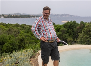Mann mit Karohemd und kurzer Hose steht an einem Pool. Im Hintergrund sind Bäume, ein größeres Gewässer und Berge zu sehen.