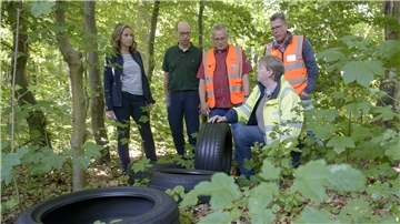 Fünf Personen, darunter eine Frau und vier Männer, stehen und knien im Wald neben illegal entsorgten Autoreifen. Drei der Männer tragen Warnkleidung.