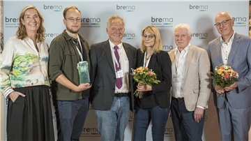 Gruppenfoto bei der Preisverleihung der Bremischen Landesmedienanstalt: Sechs Personen stehen vor einer Fotowand, drei von ihnen halten Blumensträuße oder eine Trophäe in den Händen und lächeln in die Kamera.