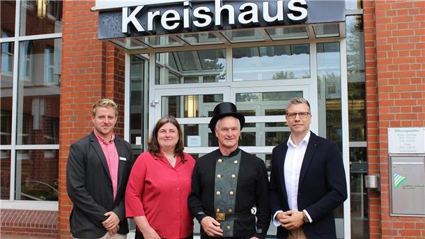 Vor dem Kreishaus in Cuxhaven stehen vier Personen nebeneinander, darunter ein Bezirksschornsteinfeger in traditioneller Uniform mit Zylinder, der seine Bestellungsurkunde erhalten hat.