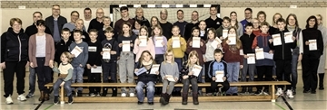Eine große Gruppe von Menschen, bestehend aus Erwachsenen und Kindern, posiert in einer Turnhalle für ein Gruppenfoto. Die Kinder in der vorderen Reihe halten Urkunden in den Händen, während die Erwachsenen dahinter stehen. Im Hintergrund ist ein Handballtor und eine helle Wand aus Klinkersteinen sichtbar.
