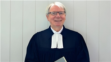 Ein evangelischer Pastor mit Talar und Brille steht vor einer Holztür.