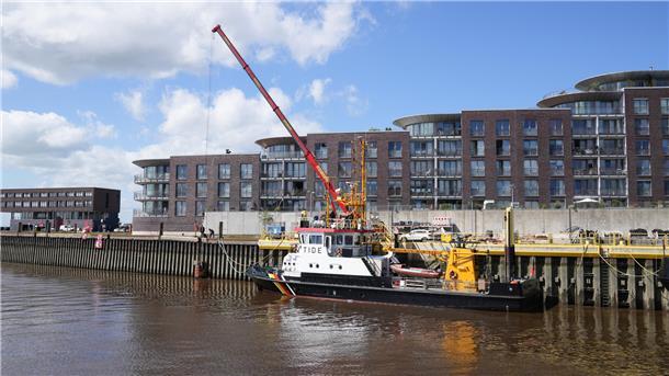 Eine Uferpromenade: An einer Kaje wird mithilfe eines Krans gearbeitet. Daneben: Ein Schiff.